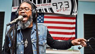 Mighty Joshua with dreadlocks singing into microphone, wearing a denim vest, in front of a US flag and a radio station banner.