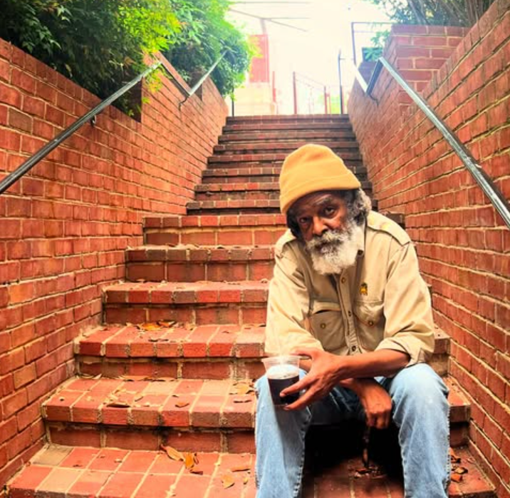 Mighty Joshua with long white beard sits on brick steps, holding a glass. He wears a yellow cap, light shirt, and jeans. Staircase ascends.