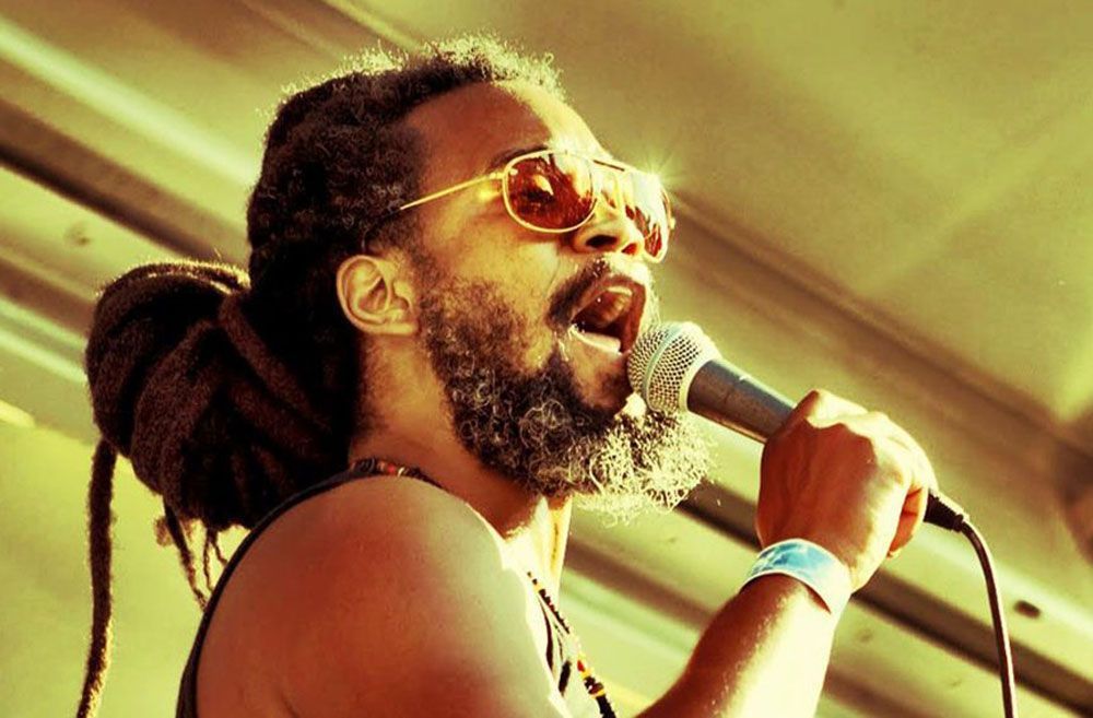 Mighty Joshua with dreadlocks and sunglasses singing into a microphone outdoors.
