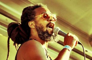 Mighty Joshua with dreadlocks singing into a microphone, wearing sunglasses, outdoors.