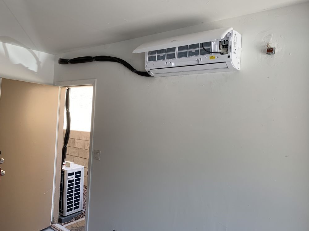 White air conditioning unit mounted on a wall with black tubing extending to an outdoor unit. Door visible.