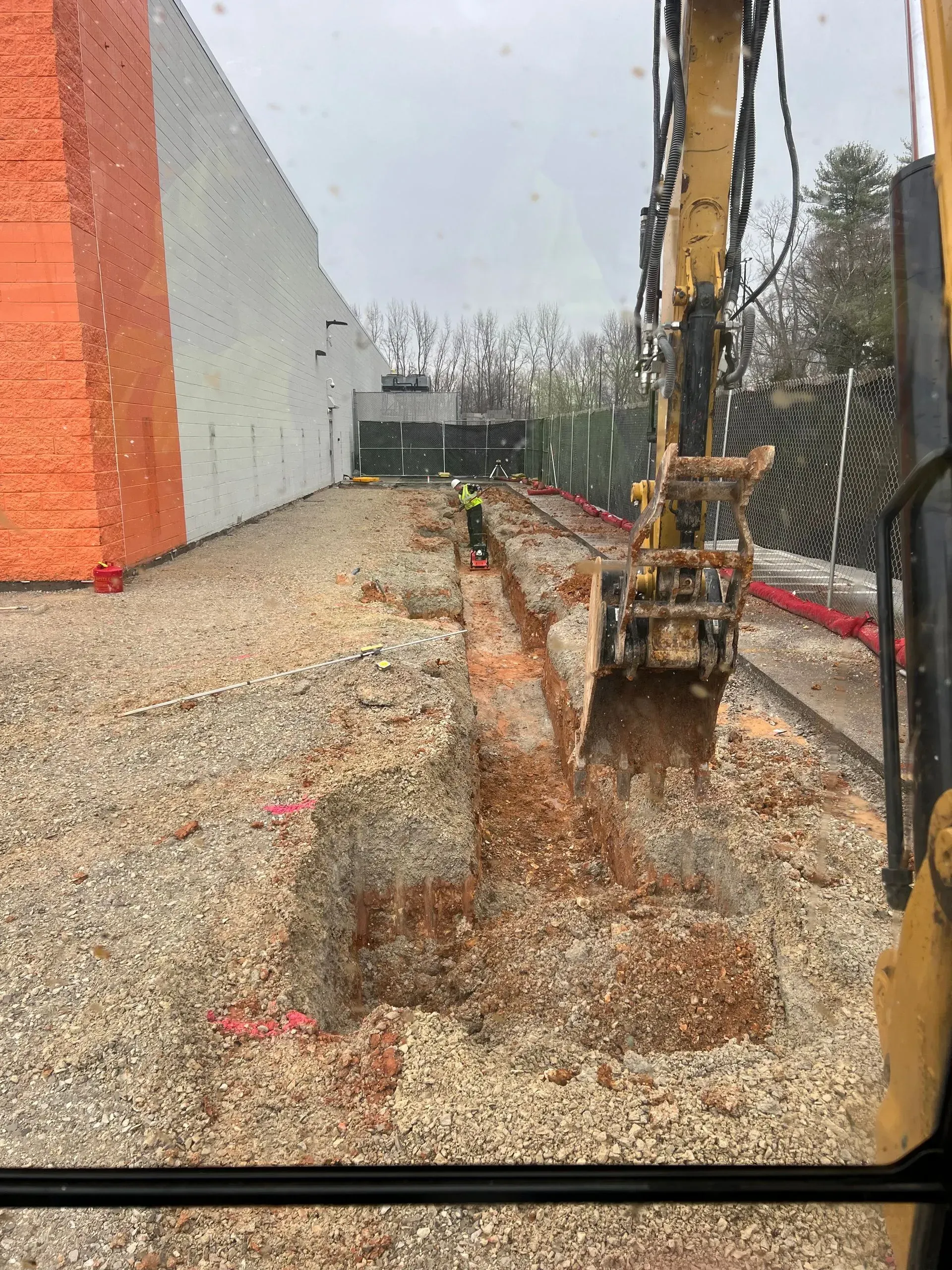 An excavator digs a trench in a gravel lot next to a building with a tall orange wall.
