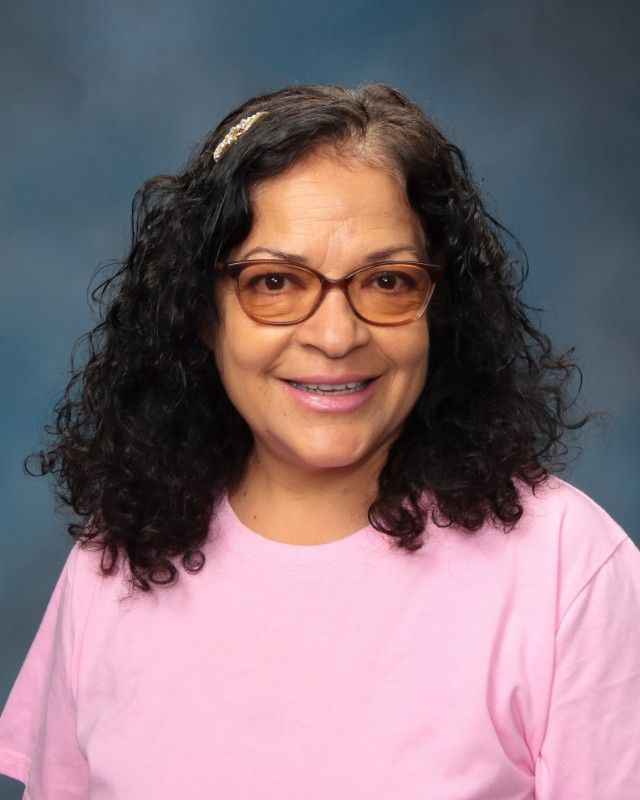 Woman with curly dark hair, wearing glasses and a pink shirt, smiles.