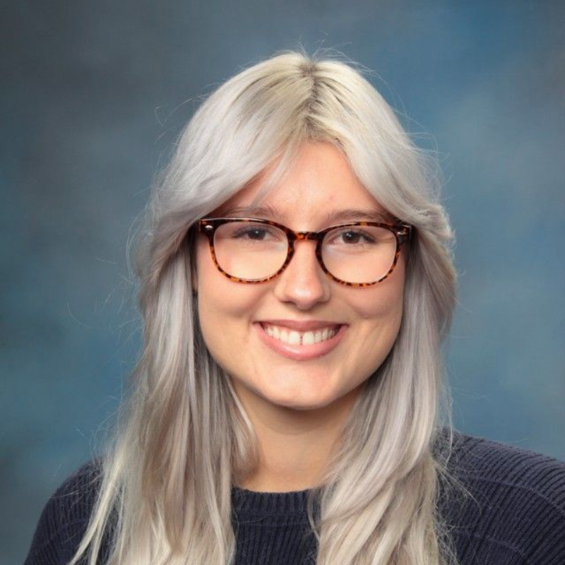 Woman with glasses smiles; light hair, blue sweater, blue background.