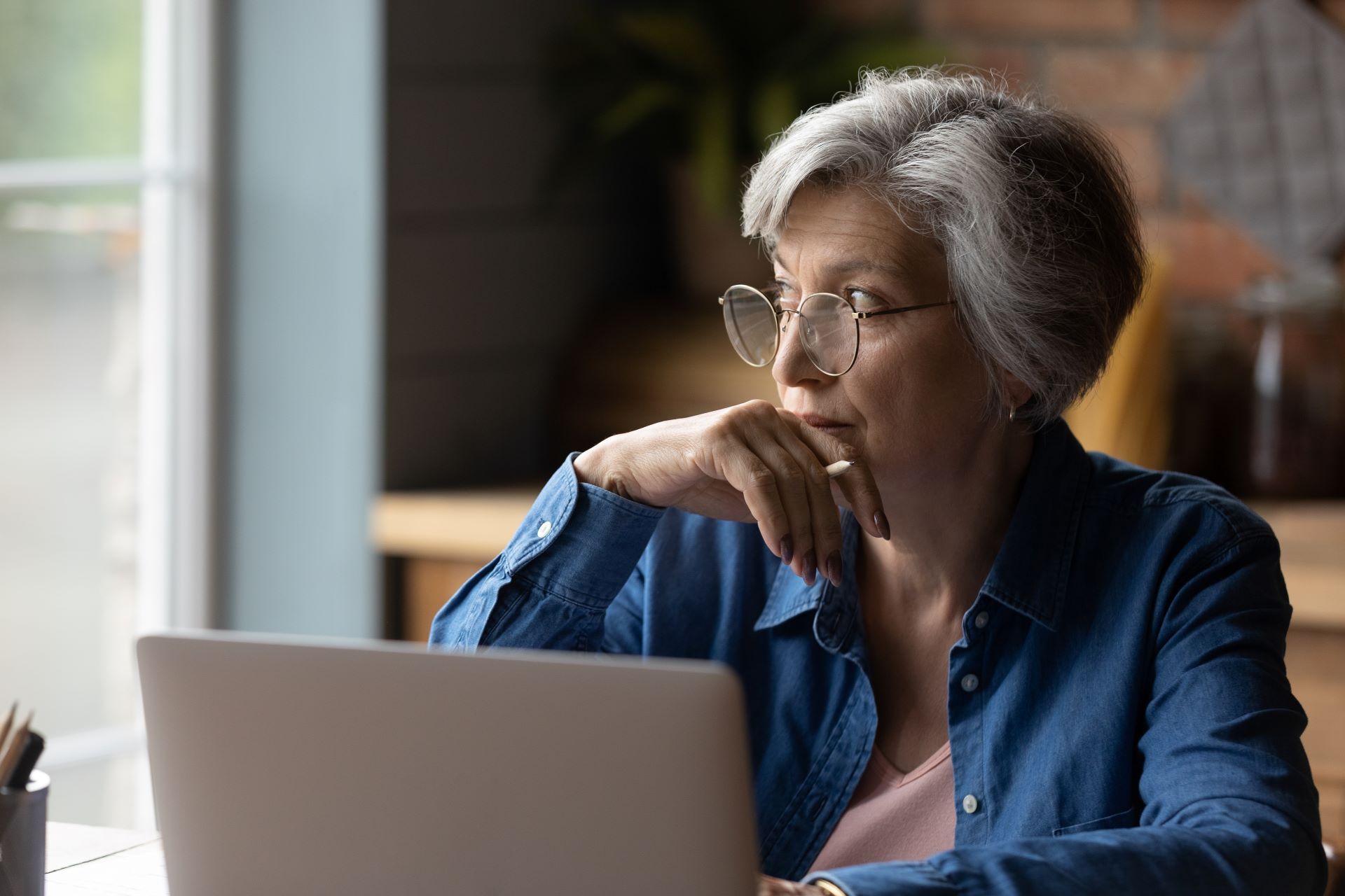 A person with gray hair and glasses wearing a denim shirt sits at a laptop, looking thoughtful while resting a hand on chin.