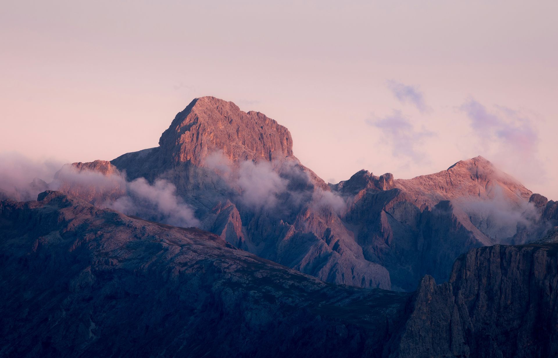 Mountains at dawn, with rocky peaks and wispy clouds under a pink and purple sky.