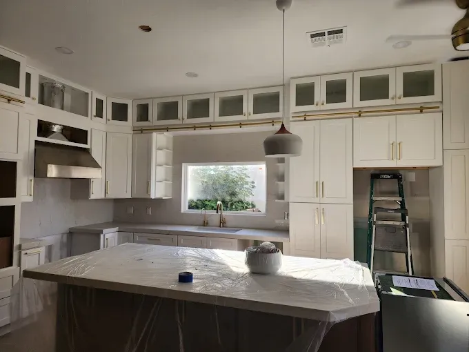 Kitchen with white cabinets, light-colored countertops, island, and window. Ladder leaning against cabinets.