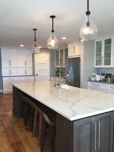 Spacious kitchen with a large island, pendant lights, and wooden floors. White cabinets and a marble countertop.