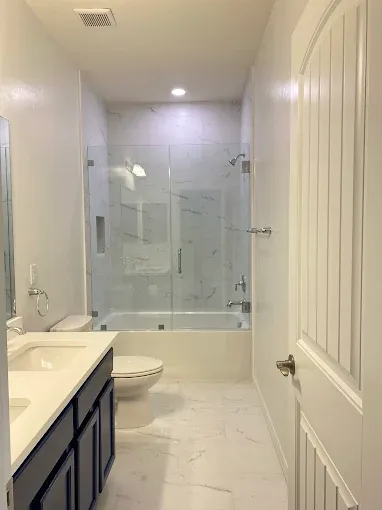 Modern bathroom with white and gray marble tile, glass shower, white cabinets, and a toilet.