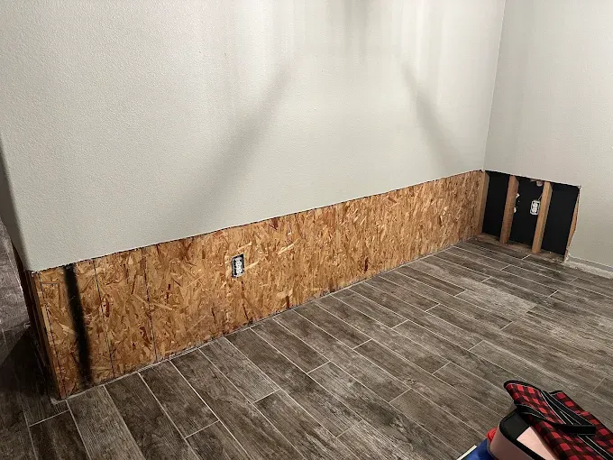 OSB paneling installed on the lower portion of a room's walls. Dark wood-look tile flooring.  An electrical outlet is visible.