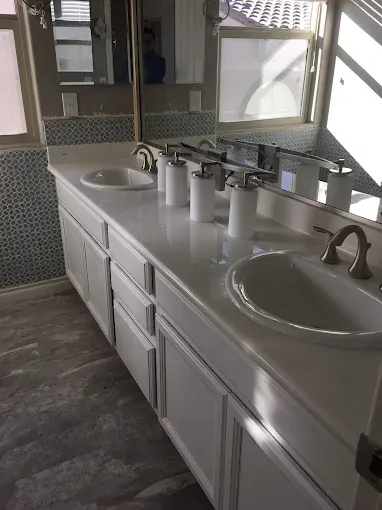 White bathroom vanity with two sinks, countertop, and mirror. White cabinetry, blue-tiled wall, and gray flooring.