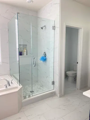 Modern bathroom with glass shower, marble tile, and a toilet visible in the doorway.