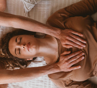 Woman receiving chest massage, eyes closed, on a bed. Two hands on her chest, neutral lighting.