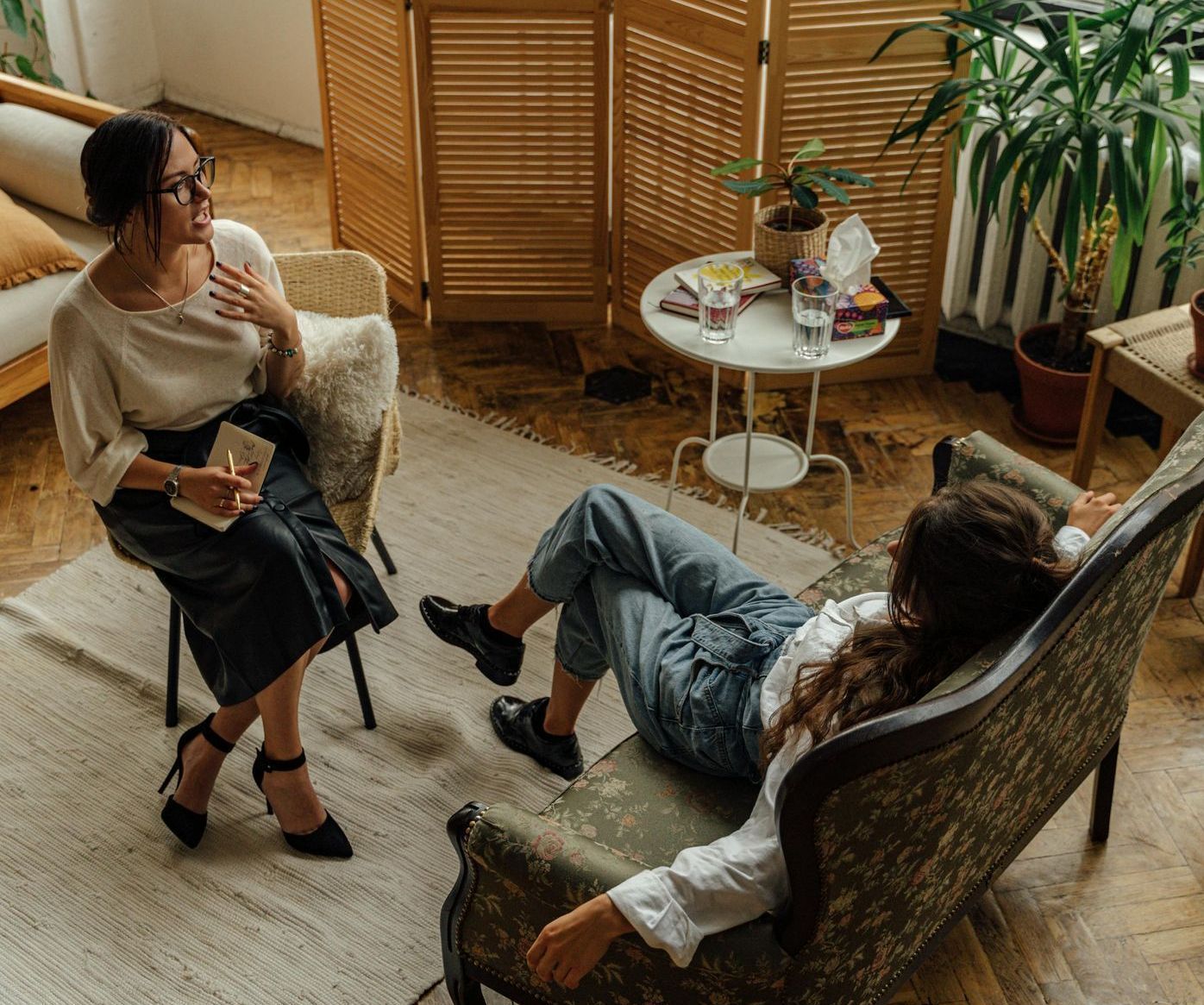 A woman is sitting in a chair talking to another woman who is laying on a couch.