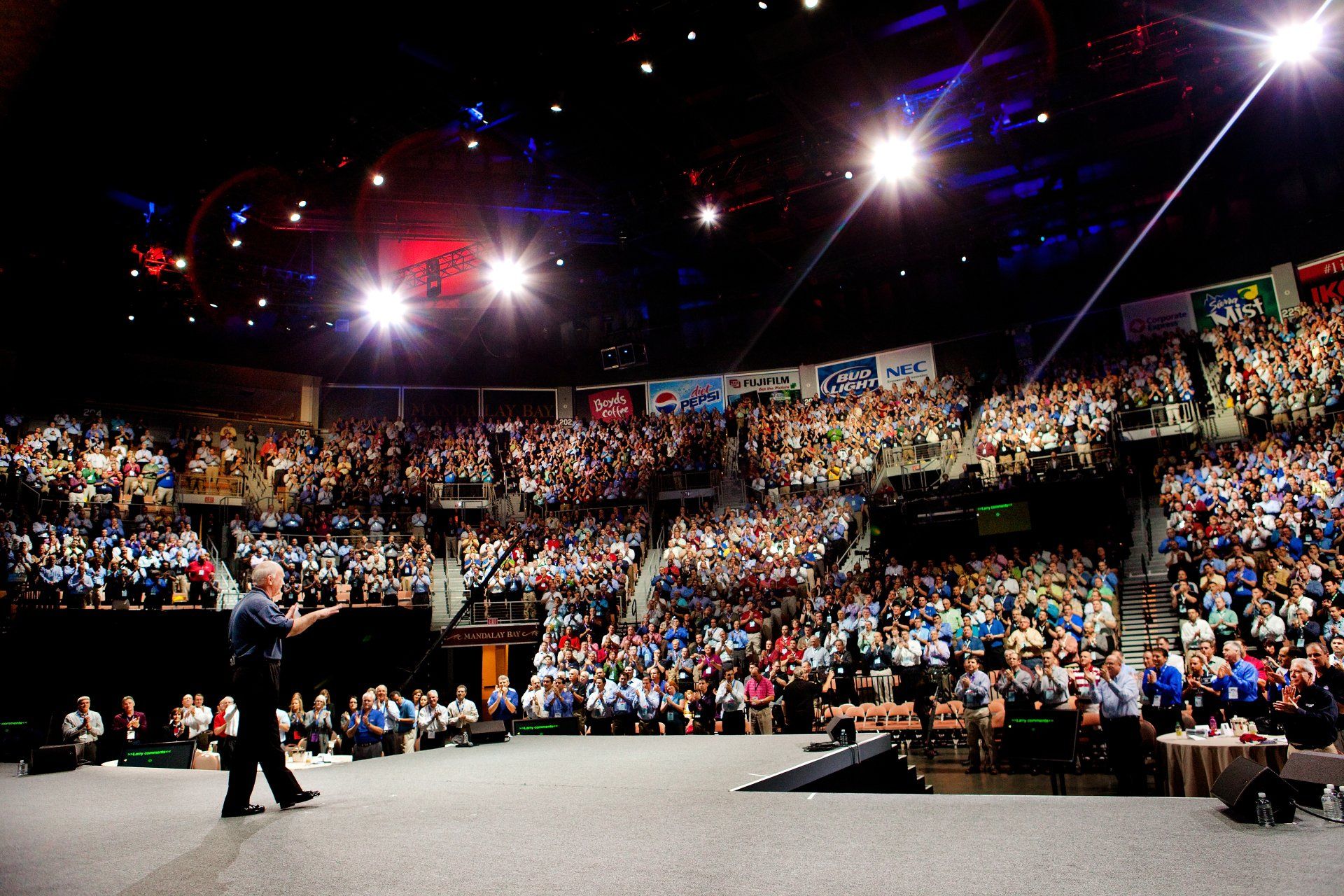 A speaker is on stage at a large corporate event. Many people are in the audience. 
