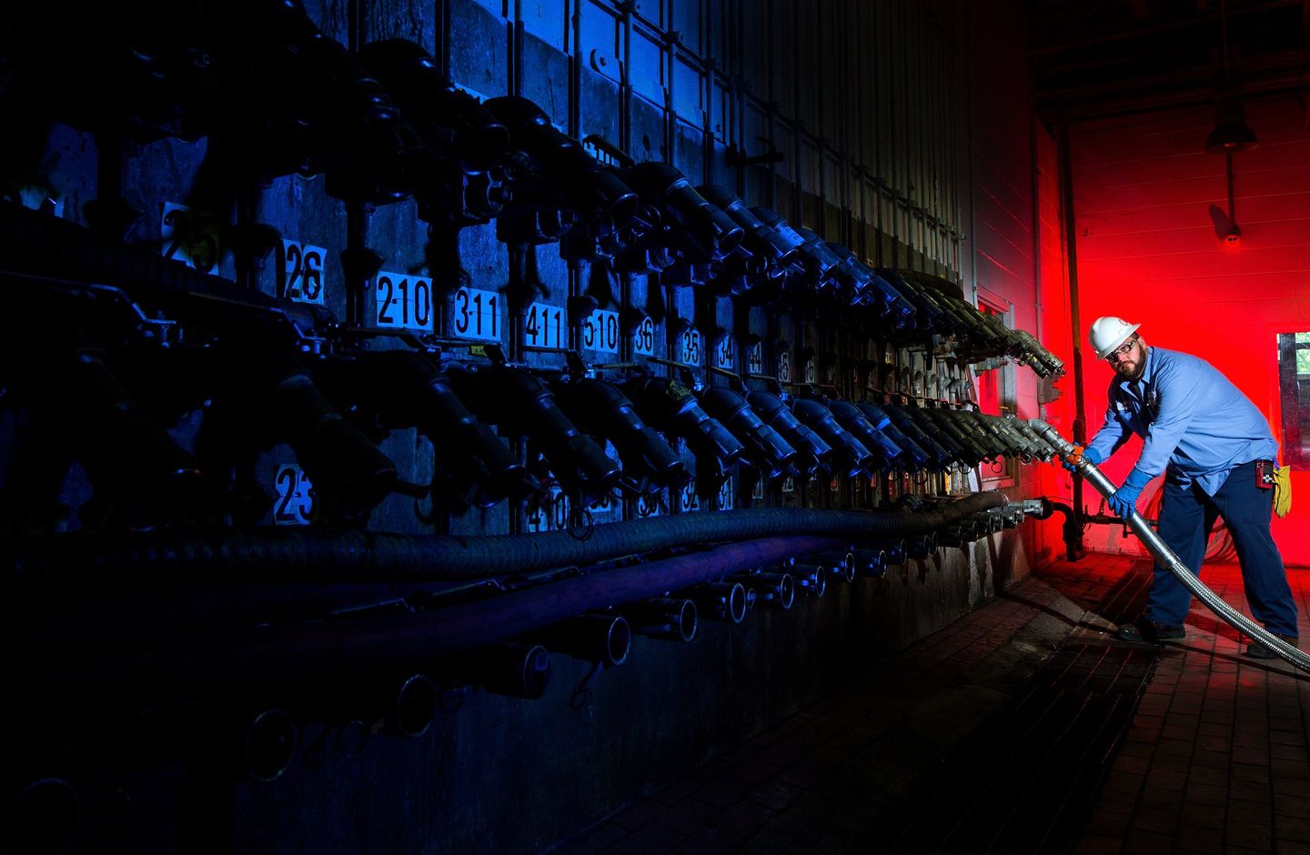 A male employee at a manufacturing facility looks at the camera as he connects a hose to a bank of connections. He is wearing safety gear. 