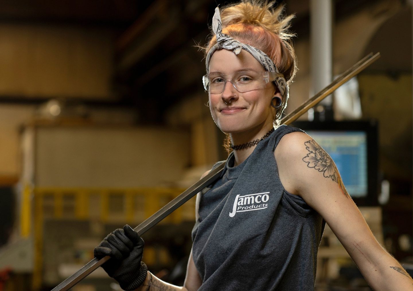 A woman who works in a manufacturing facility poses for the camera. She is holding a rod in her gloved hand and is looking at the camera. She is wearing safety glasses. 
