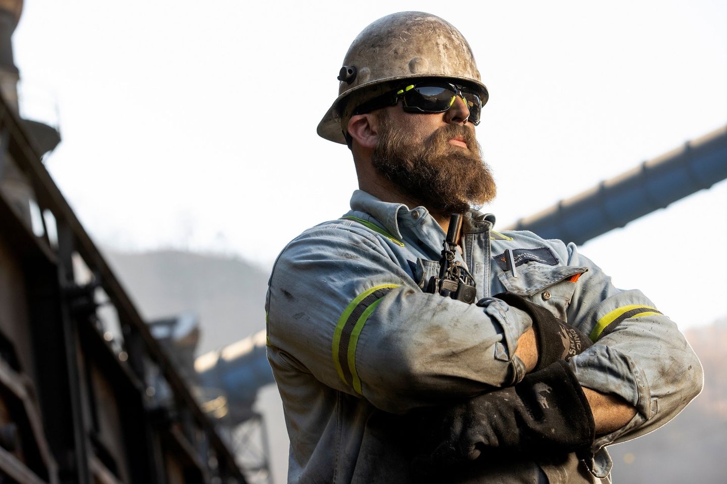 A male industrial plant employee poses to have his photo taken outside of the plant. The plant is visible behind him. He is wearing safety gear and his clothing is dirty from work. 