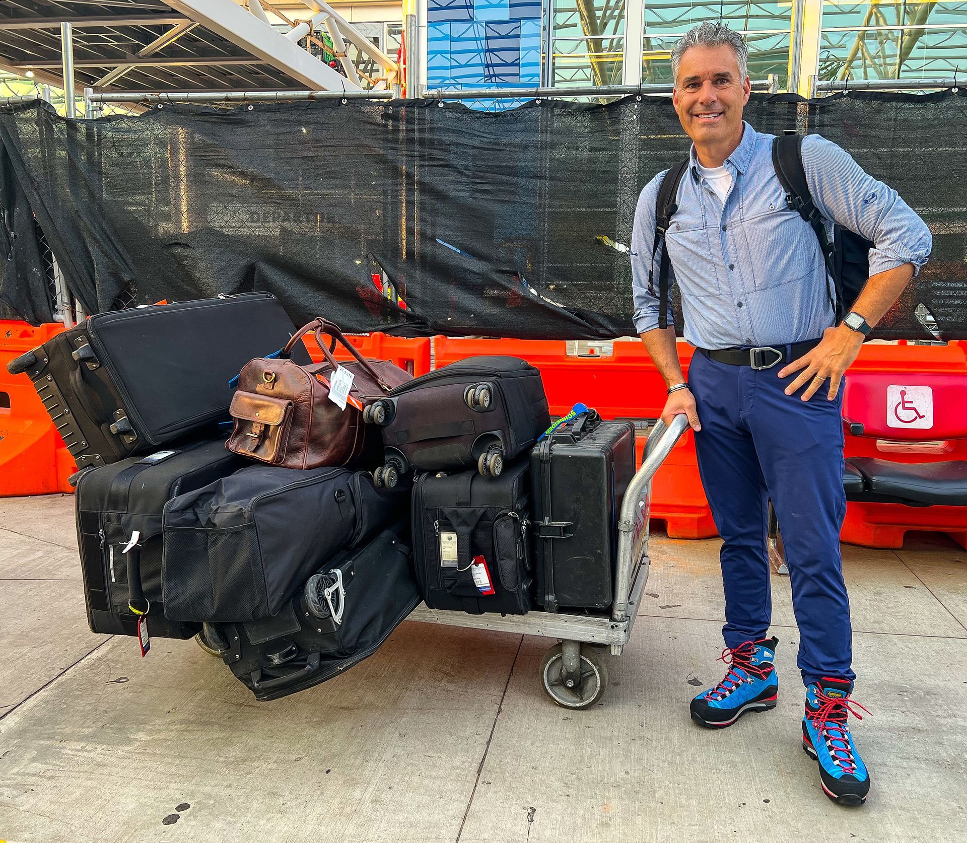 Man standing next to luggage cart that is filled with large bags. He is at airport.