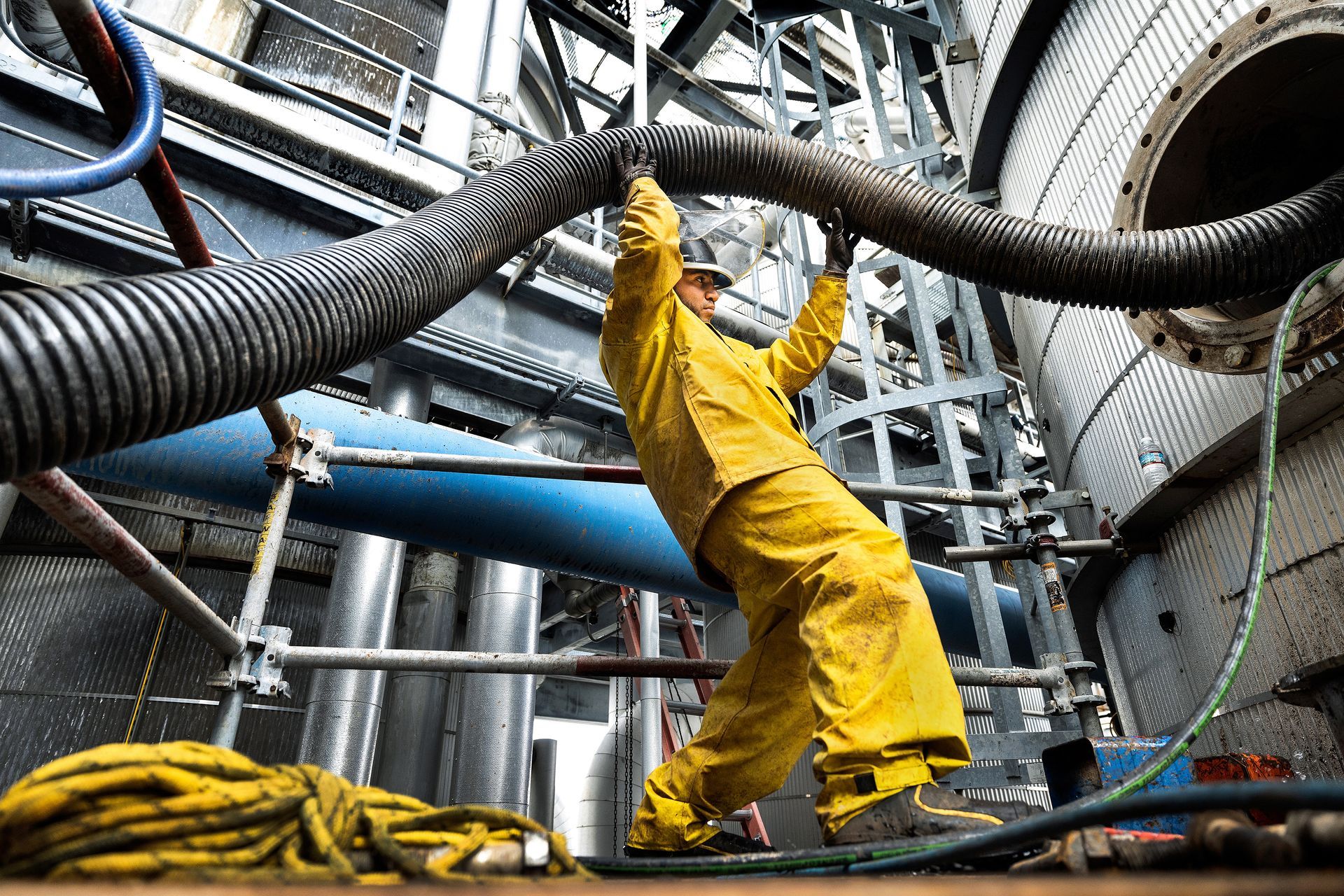 Photography of an employee performing industrial cleaning at a gas refinery. The male employee is wearing a bright yellow rain suit and is holding a large hose over his head. 