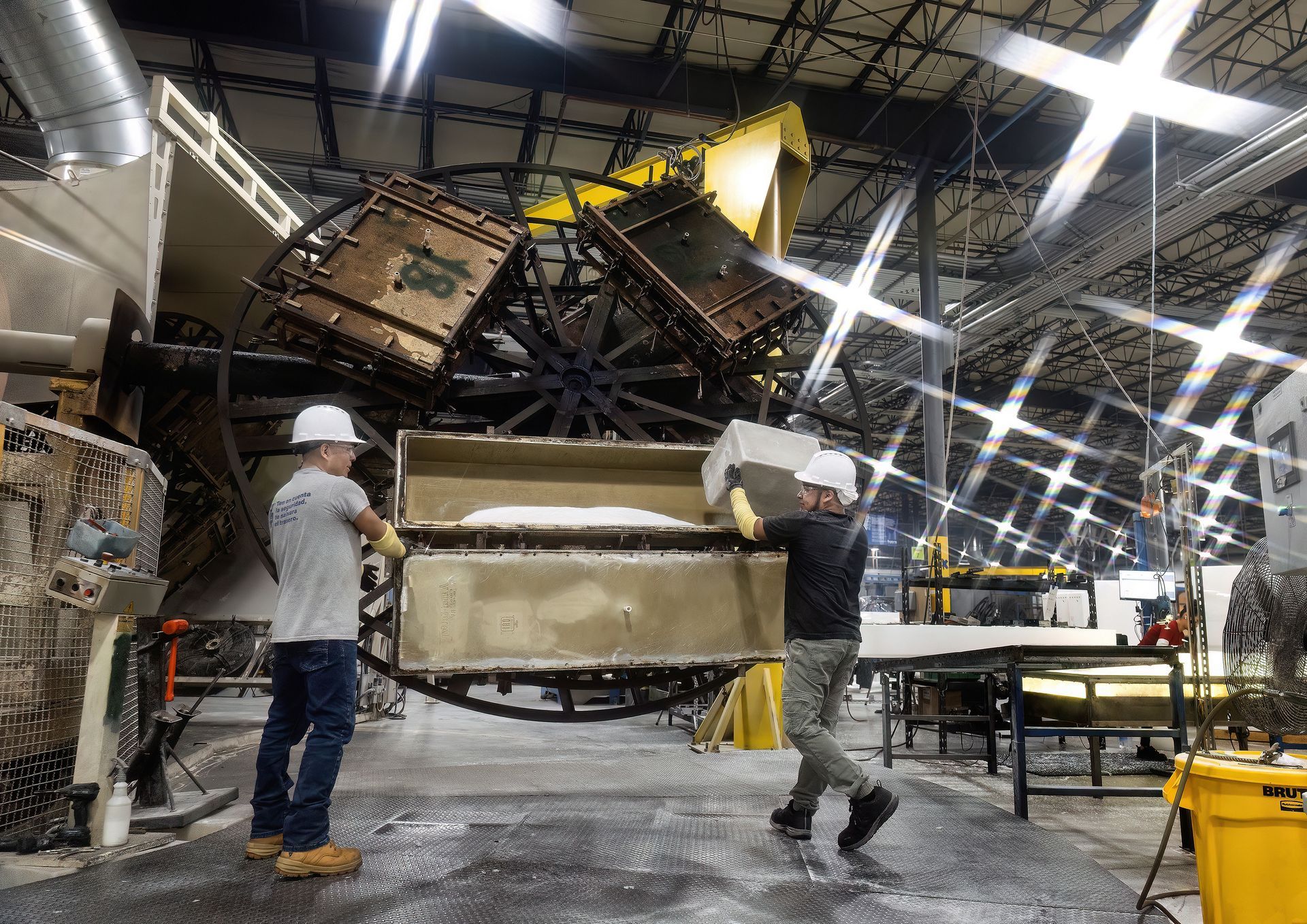 Two male workers in a plastics molding company dump white plastic pellets into a hopper. The men are wearing protective safety equipment and are using a large bin to transport the pellets to the hopper. 