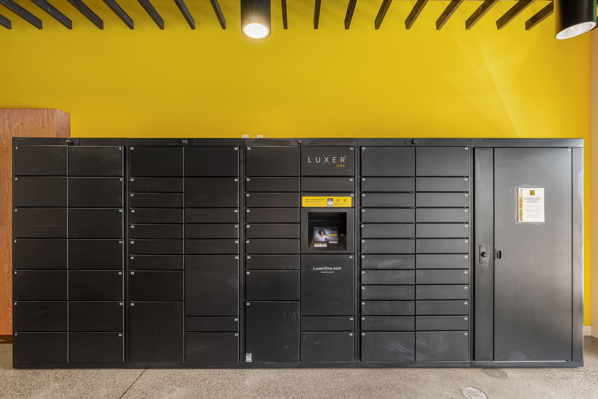 Black parcel locker wall with central touchscreen kiosk under a yellow ceiling indoors at Anthem on 12th, offering luxury apartments in Capitol Hill.
