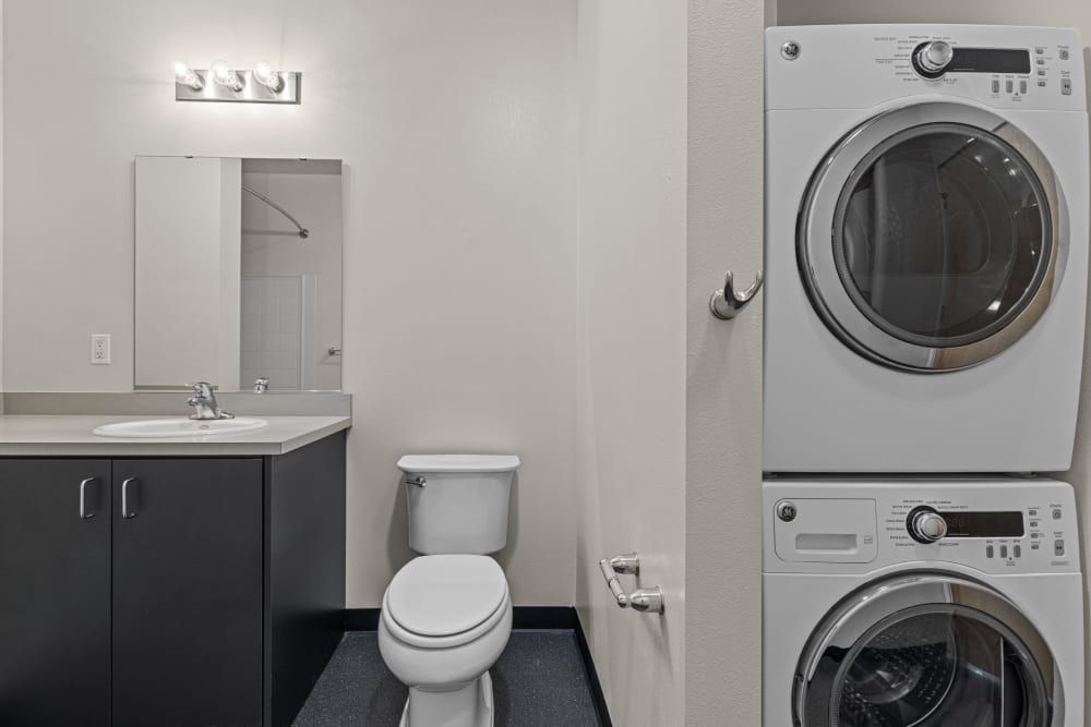 A modern, compact bathroom with a combination of laundry appliances. To the right, a stacked washer and dryer set in white, and to the left, a bathroom vanity with a black base and a white countertop featuring a sink and a faucet. Above the vanity is a rectangular mirror flanked by a four-lamp light fixture. A white toilet is situated next to the vanity, and the room is painted in a light neutral color with dark flooring for contrast at Anthem on 12th in Seattle, WA.