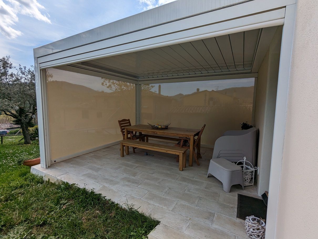 Patio coperto con tavolo e sedie in legno, pavimento in cemento e barbecue sotto una tettoia aperta su un lato.