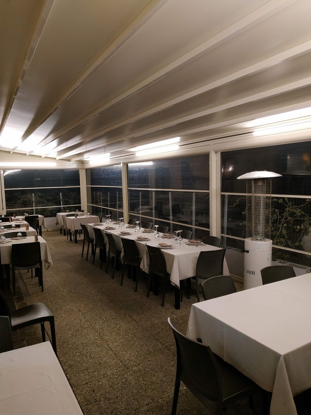 Sala da pranzo vuota di un ristorante con tovaglie bianche, sedie nere e grandi finestre, di notte.