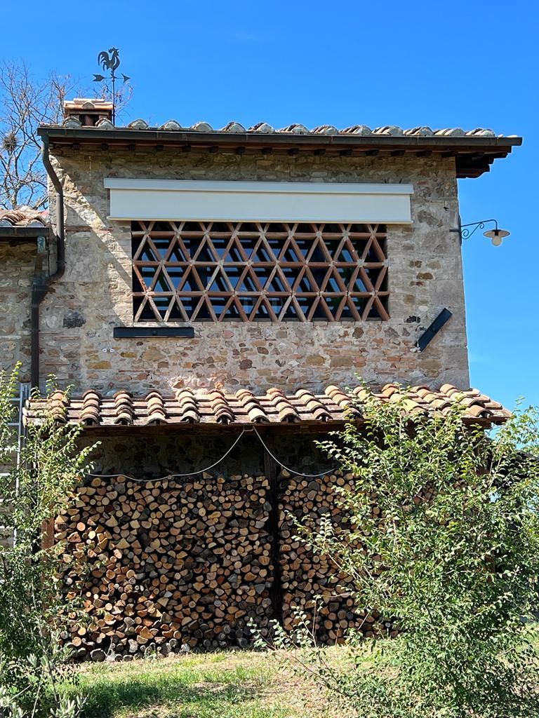 Casa rustica in pietra con tetto di tegole, finestre con grate in legno e legna da ardere accatastata sotto un cielo azzurro.
