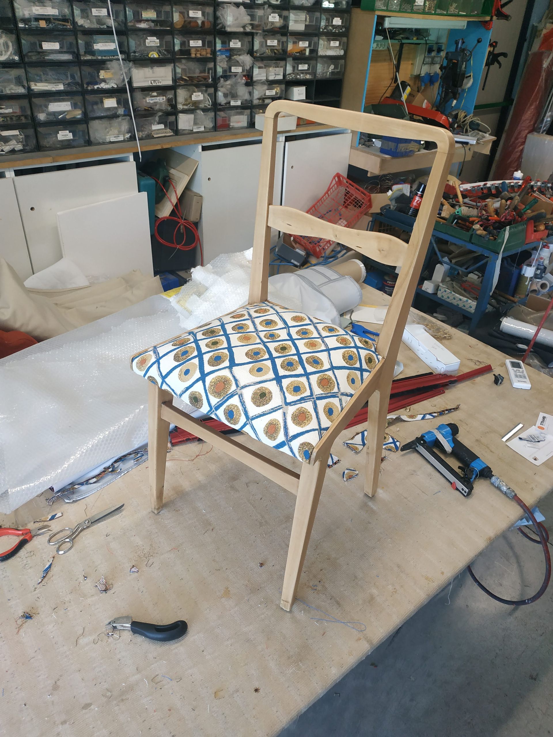 Struttura di sedia in legno con cuscino decorato in un'officina disordinata, circondata da attrezzi e materiali.