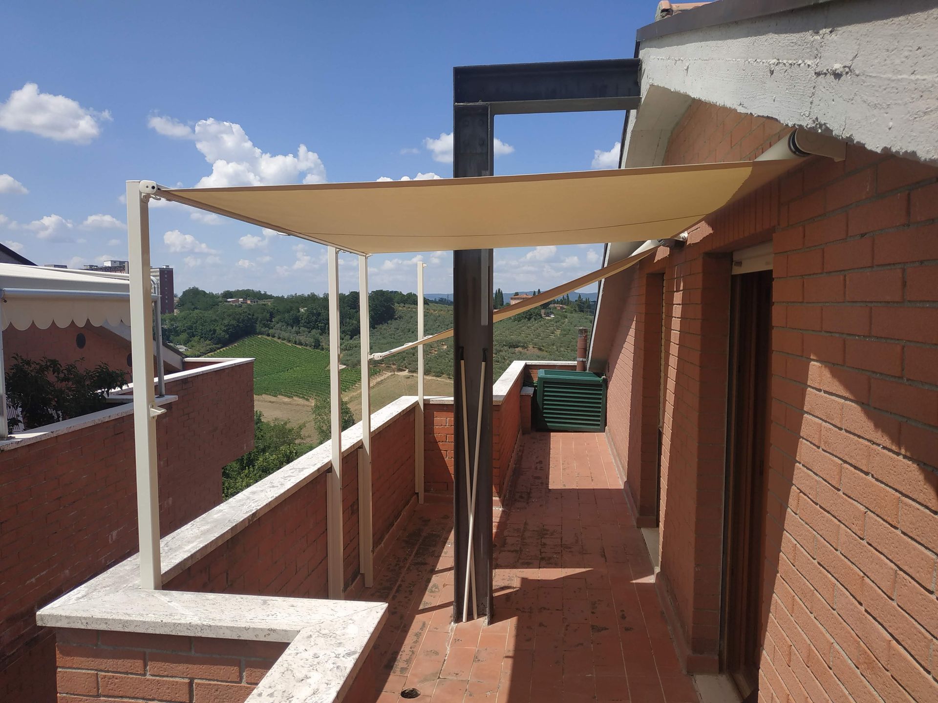 Terrazza sul tetto con tenda parasole beige, muri in mattoni e vista su verdi colline sotto un cielo azzurro.