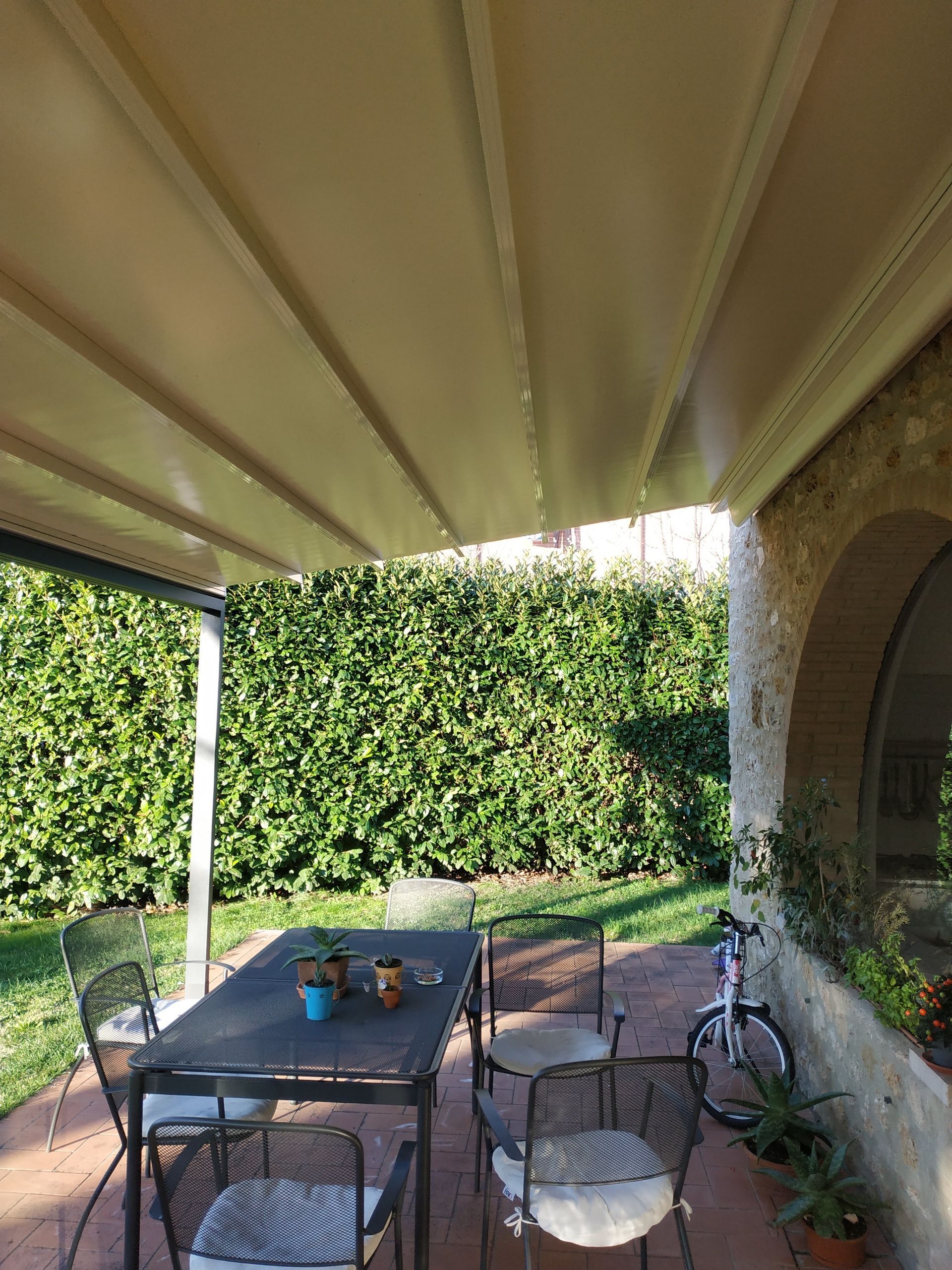 Patio con tavolo, sedie, bicicletta e fiori in vaso sotto una tenda da sole beige accanto a un'alta siepe.