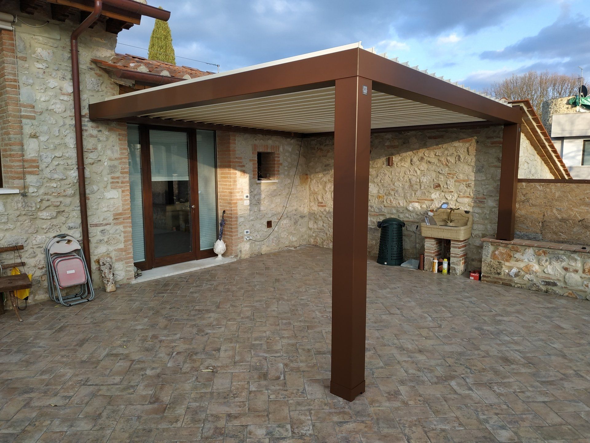 Patio in pietra con pergolato marrone accanto a una casa rustica e zona salotto esterna.