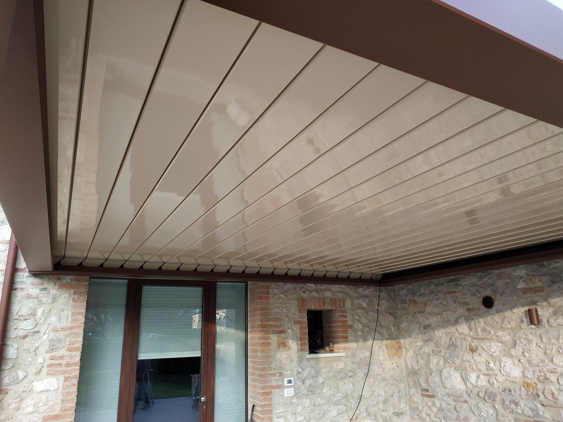 Tettoia del patio con muro in pietra, porta scorrevole in vetro e piccola finestra sotto la sporgenza del tetto della casa.