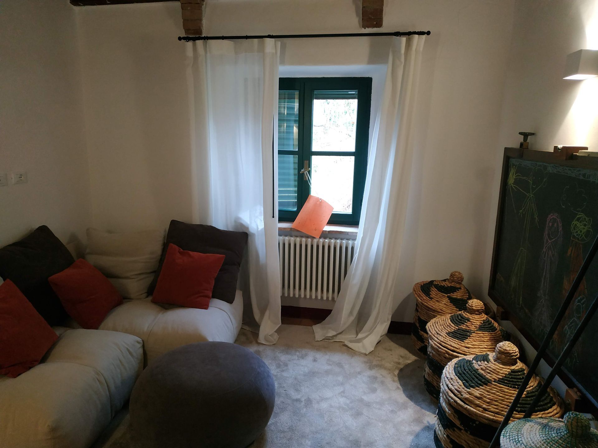 Accogliente soggiorno con divano, cuscini, pouf rotondo, tende trasparenti e sedile vicino a una piccola finestra.