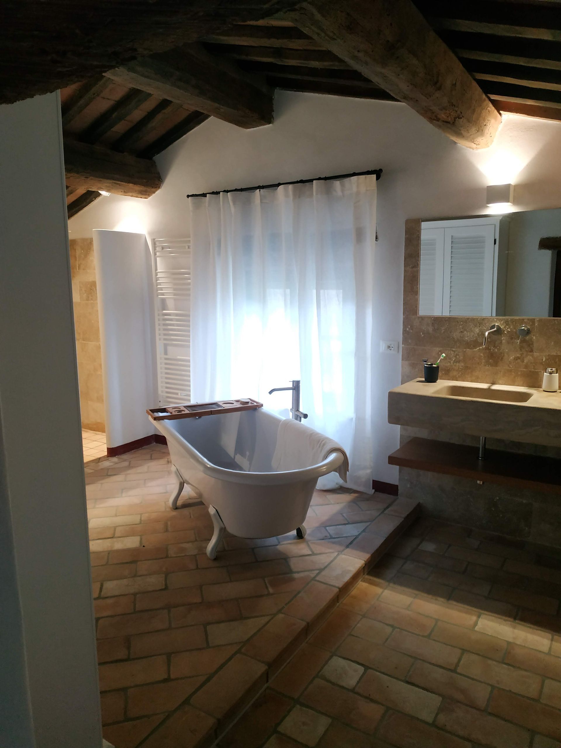 Bagno rustico con vasca freestanding, pavimento piastrellato, travi a vista e mobile lavabo accanto a una finestra.