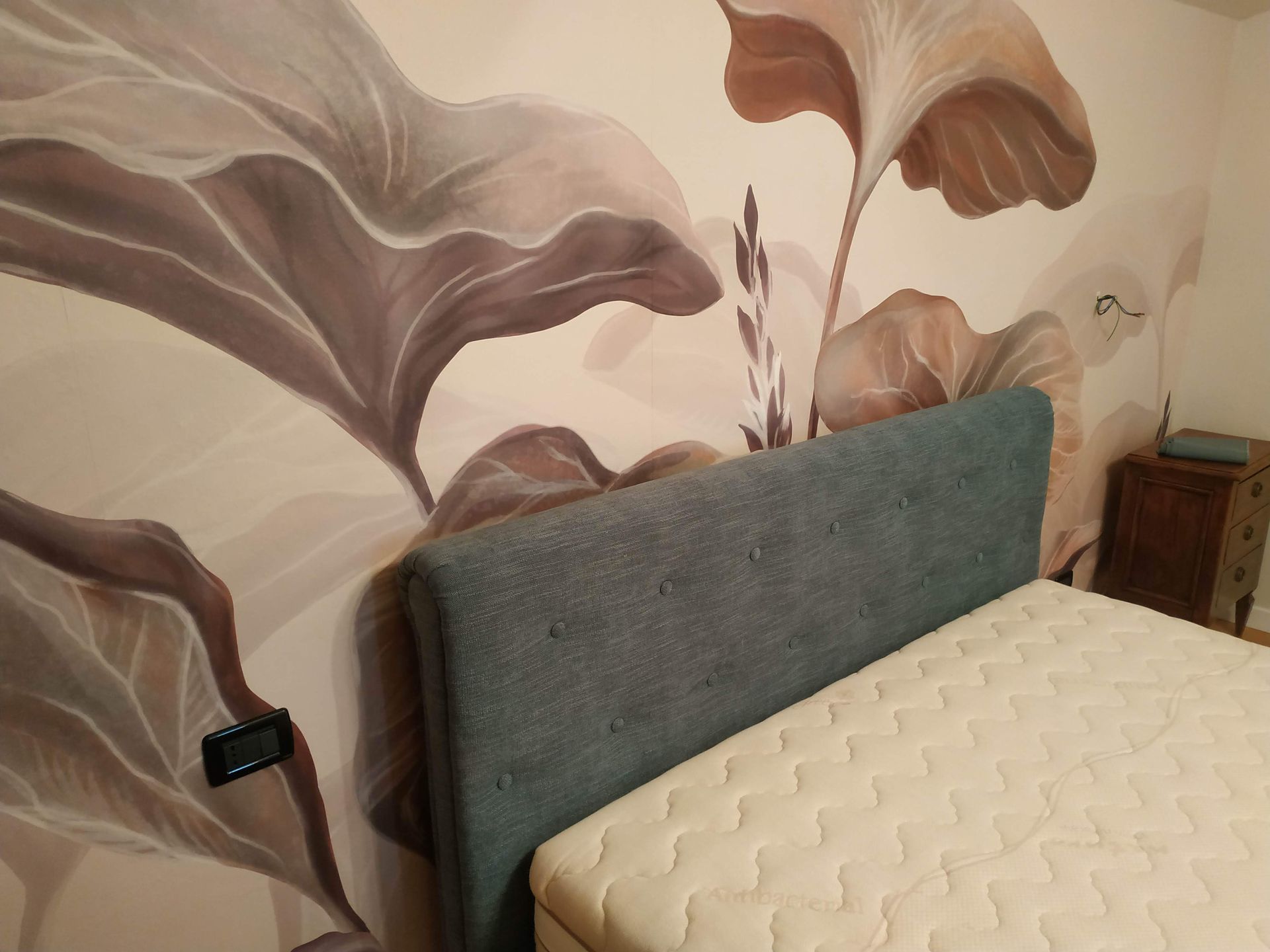 Camera da letto con testiera imbottita grigia, materasso beige e grande murale floreale sulla parete.