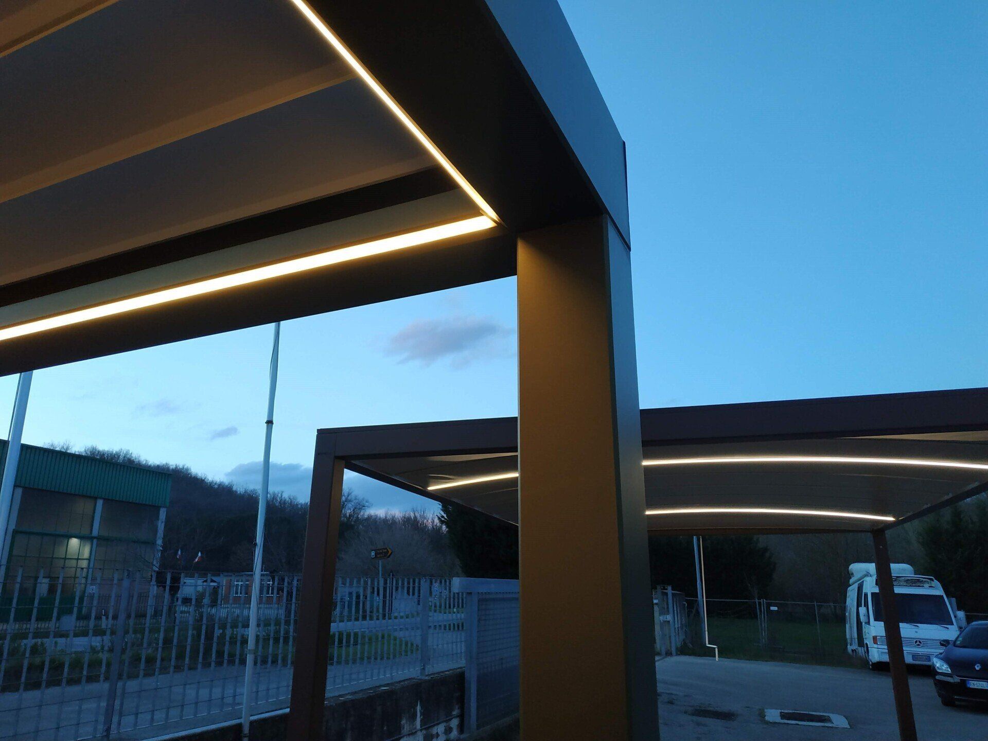 Moderna passerella esterna con strutture a pergola illuminate al crepuscolo e un furgone bianco parcheggiato sullo sfondo.