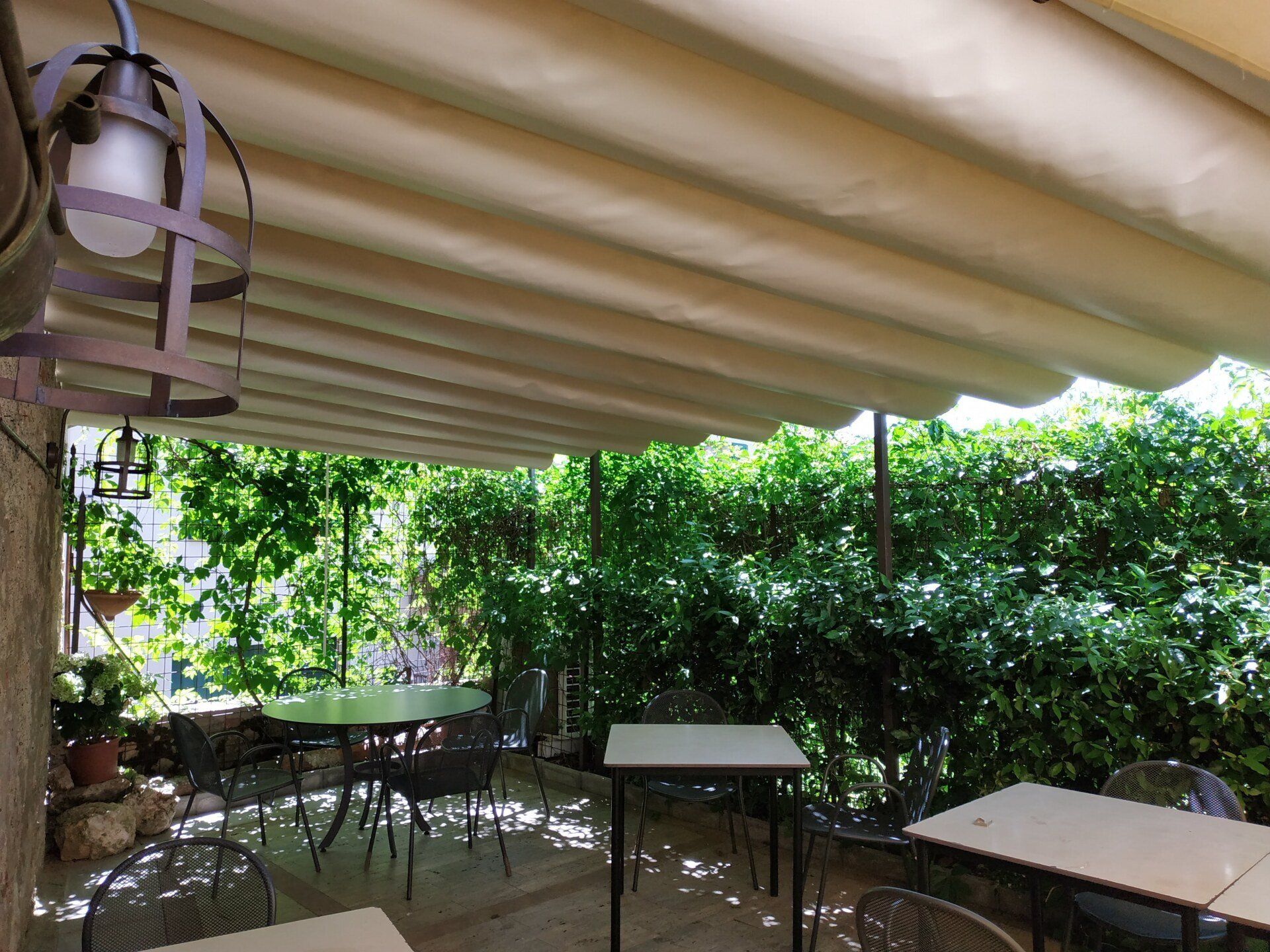 Patio esterno ombreggiato con tavoli, sedie e rigogliose siepi verdi sotto una tenda a righe.