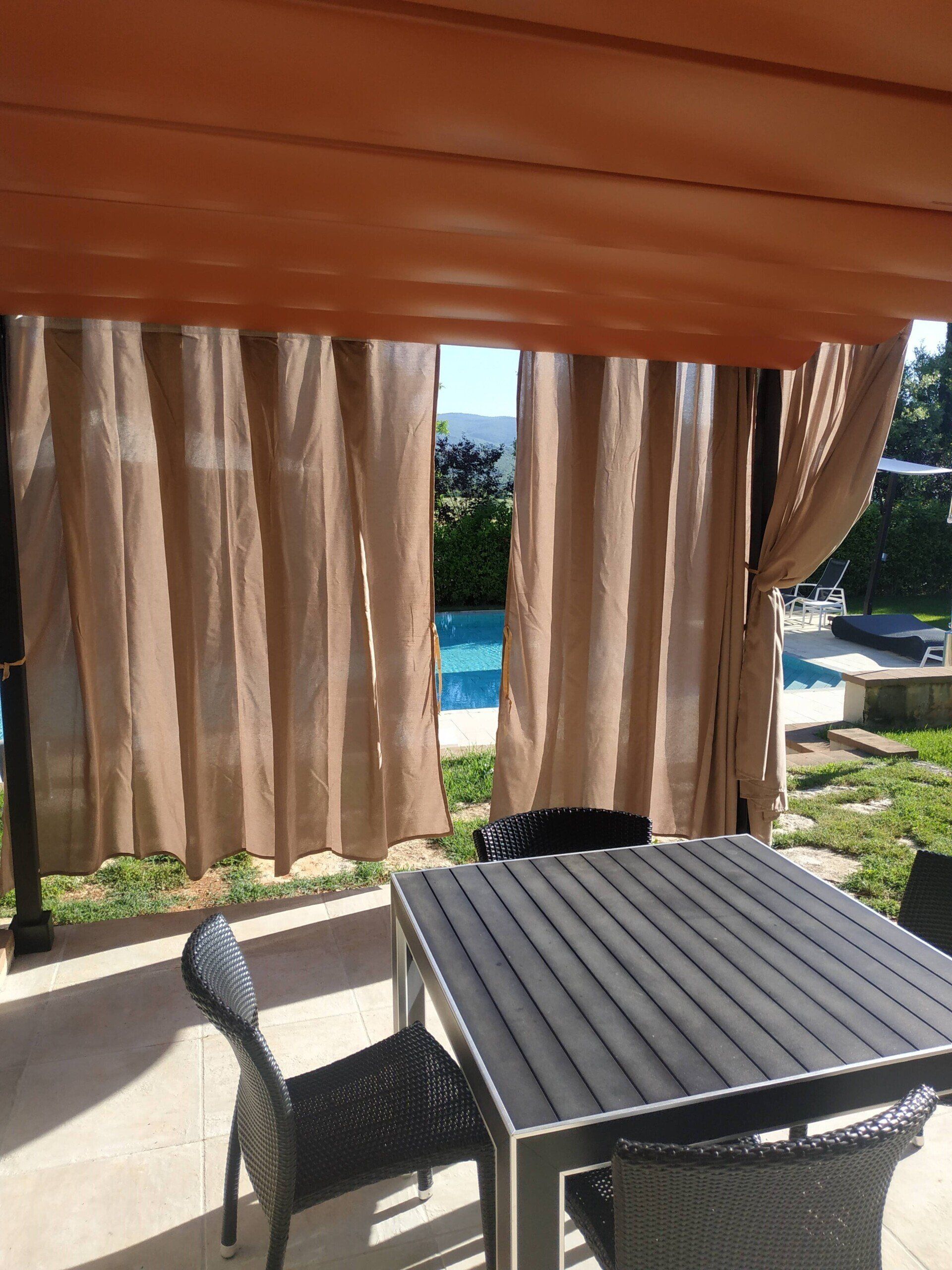Tavolo e sedia da patio accanto a tende beige, con una piscina e vegetazione sullo sfondo.