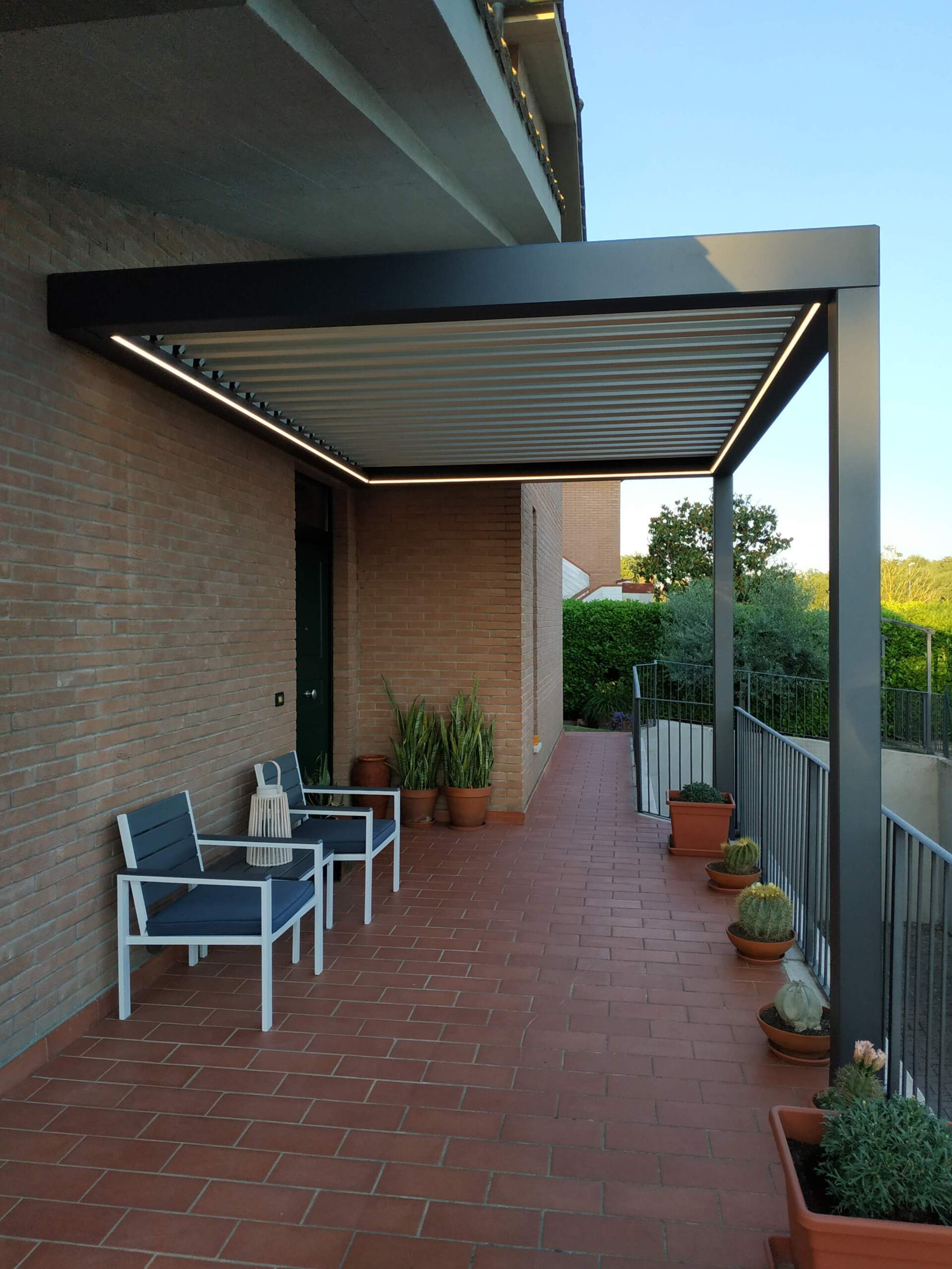 Patio coperto in mattoni con sedie in metallo, piante in vaso e tenda da sole retrattile, adiacente a una casa.