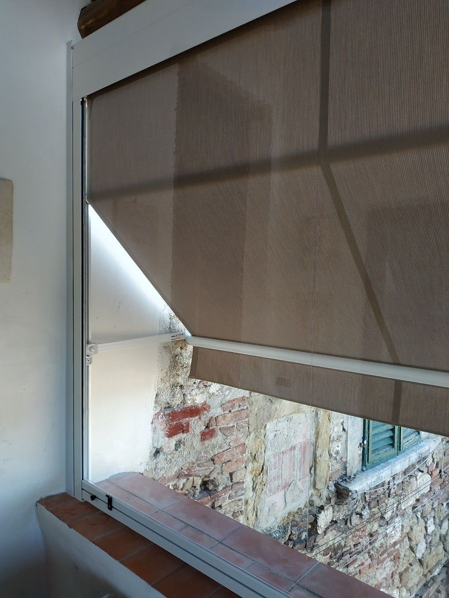 Tenda a rullo marrone parzialmente abbassata sopra un muro di pietra e un davanzale di legno, con luce naturale visibile dall'esterno.