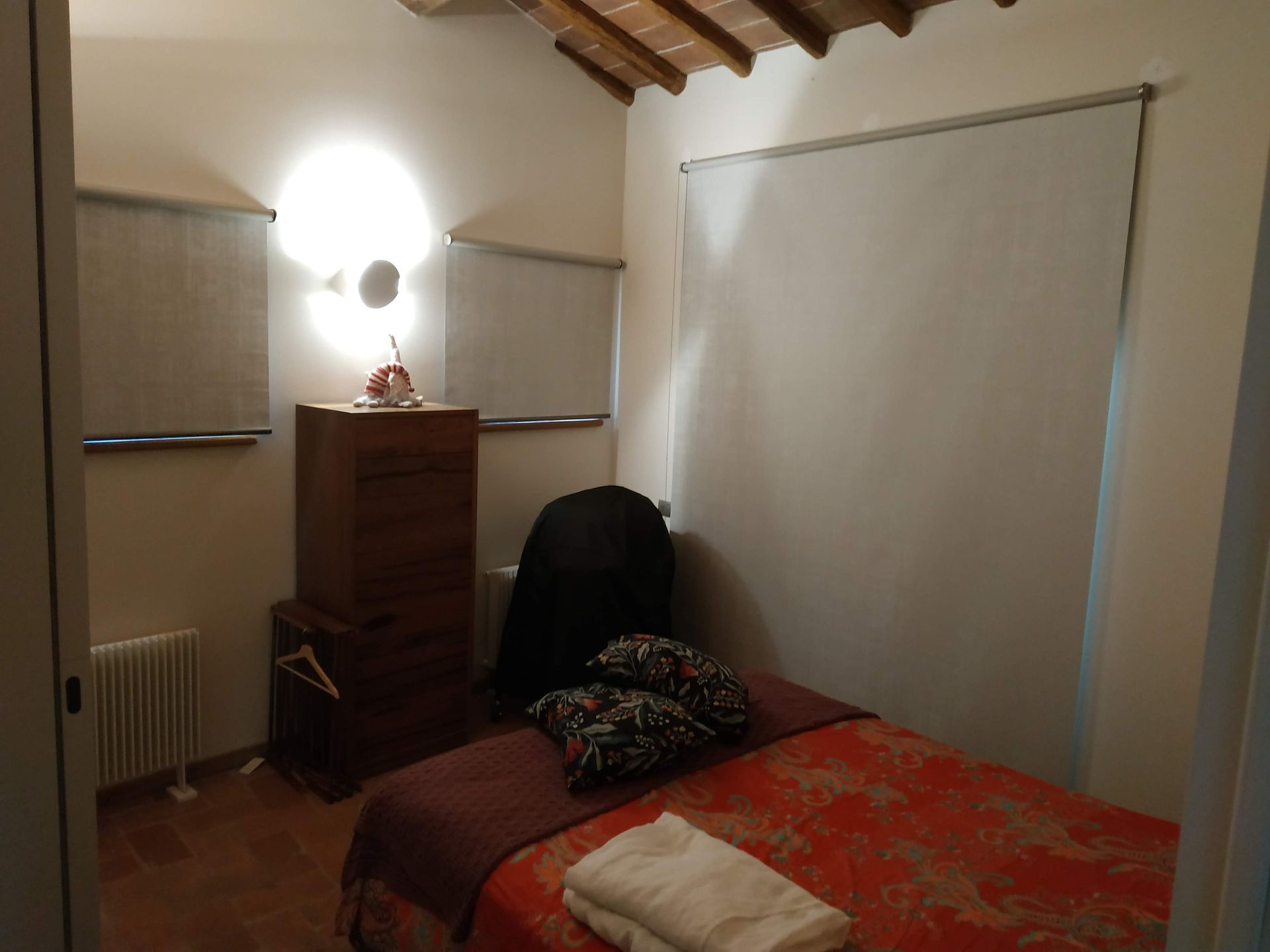 Piccola camera da letto con letto, comò, sedia, lampada e persiane chiuse sotto un soffitto di legno inclinato.