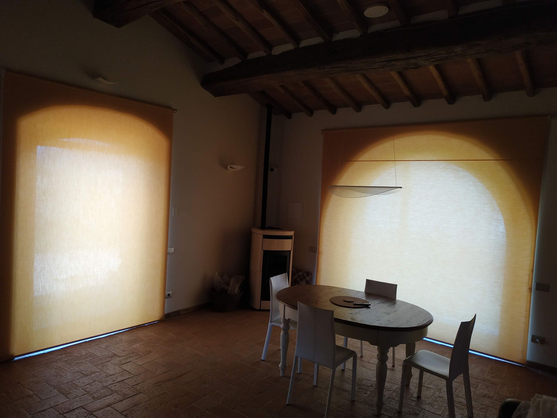 Stanza rustica vuota con soffitto in legno, pavimento piastrellato, tavolo rotondo, sedie e grandi finestre luminose.