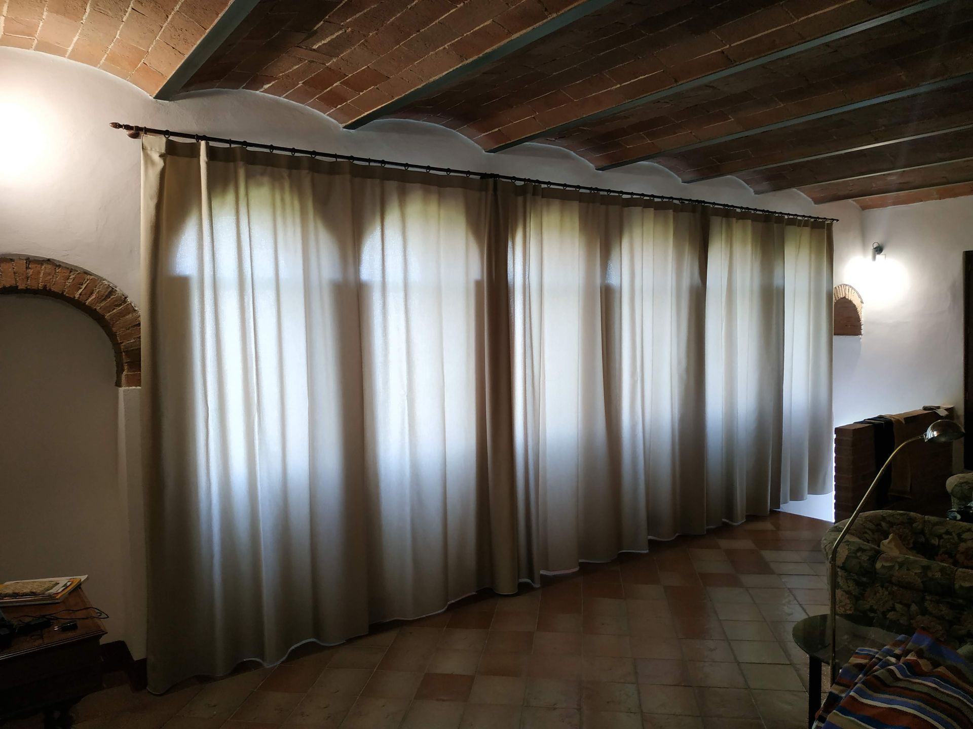 Parete finestrata con tende in una stanza rustica con pavimento piastrellato, dettagli ad arco in mattoni e soffitto con travi in ​​legno.