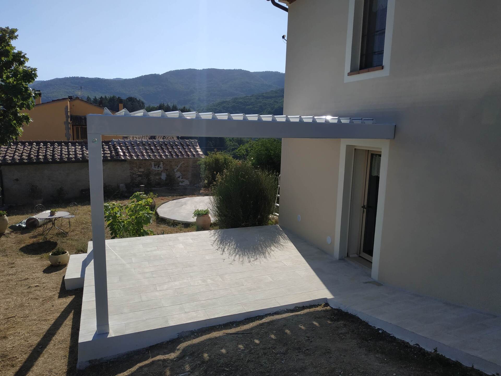 Patio di una casa moderna con pergola bianca, pavimento piastrellato e vista sulle montagne sullo sfondo.