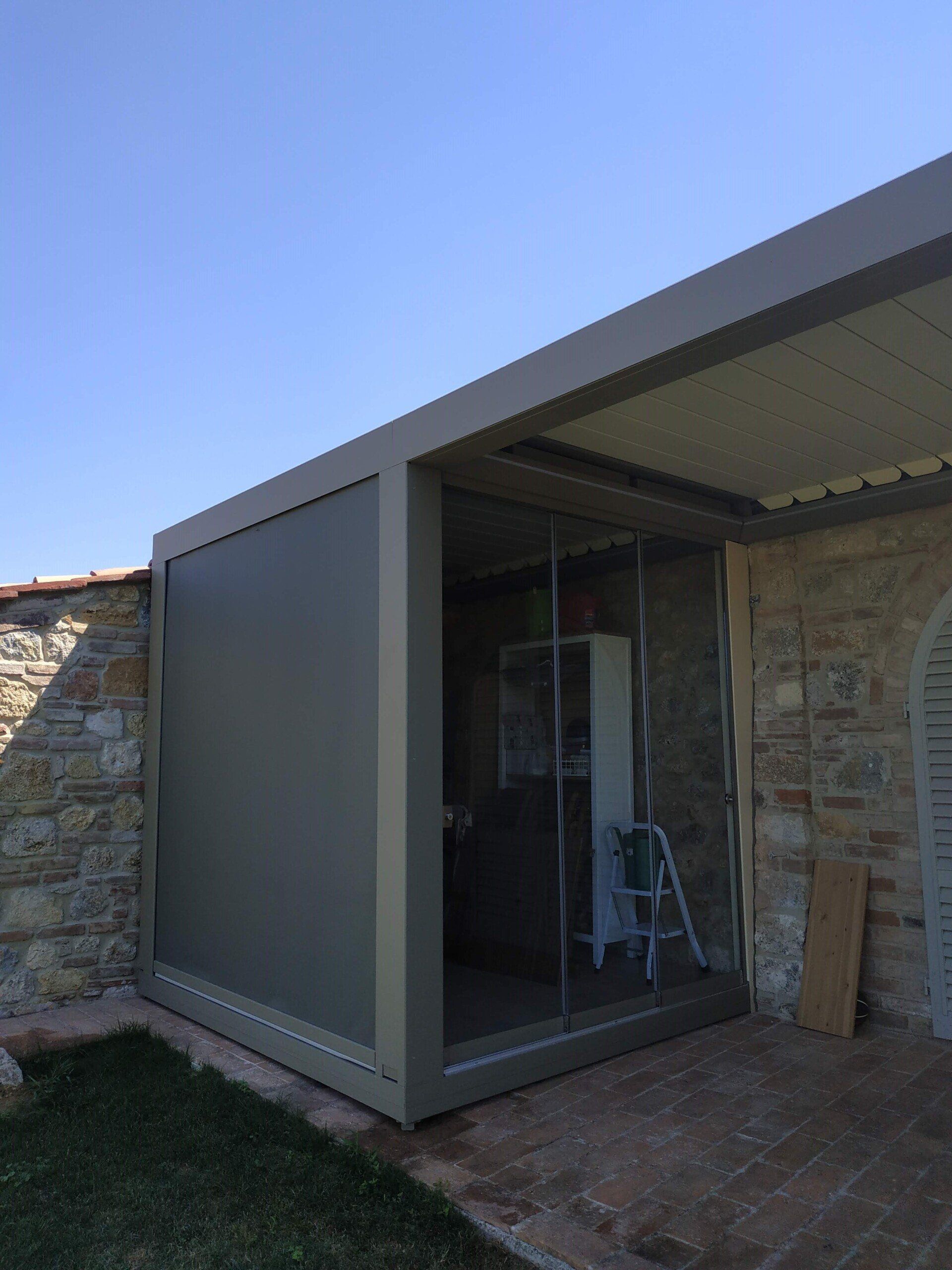 Ingresso moderno di una casa in pietra con porta scorrevole in vetro grigio e patio ombreggiato.