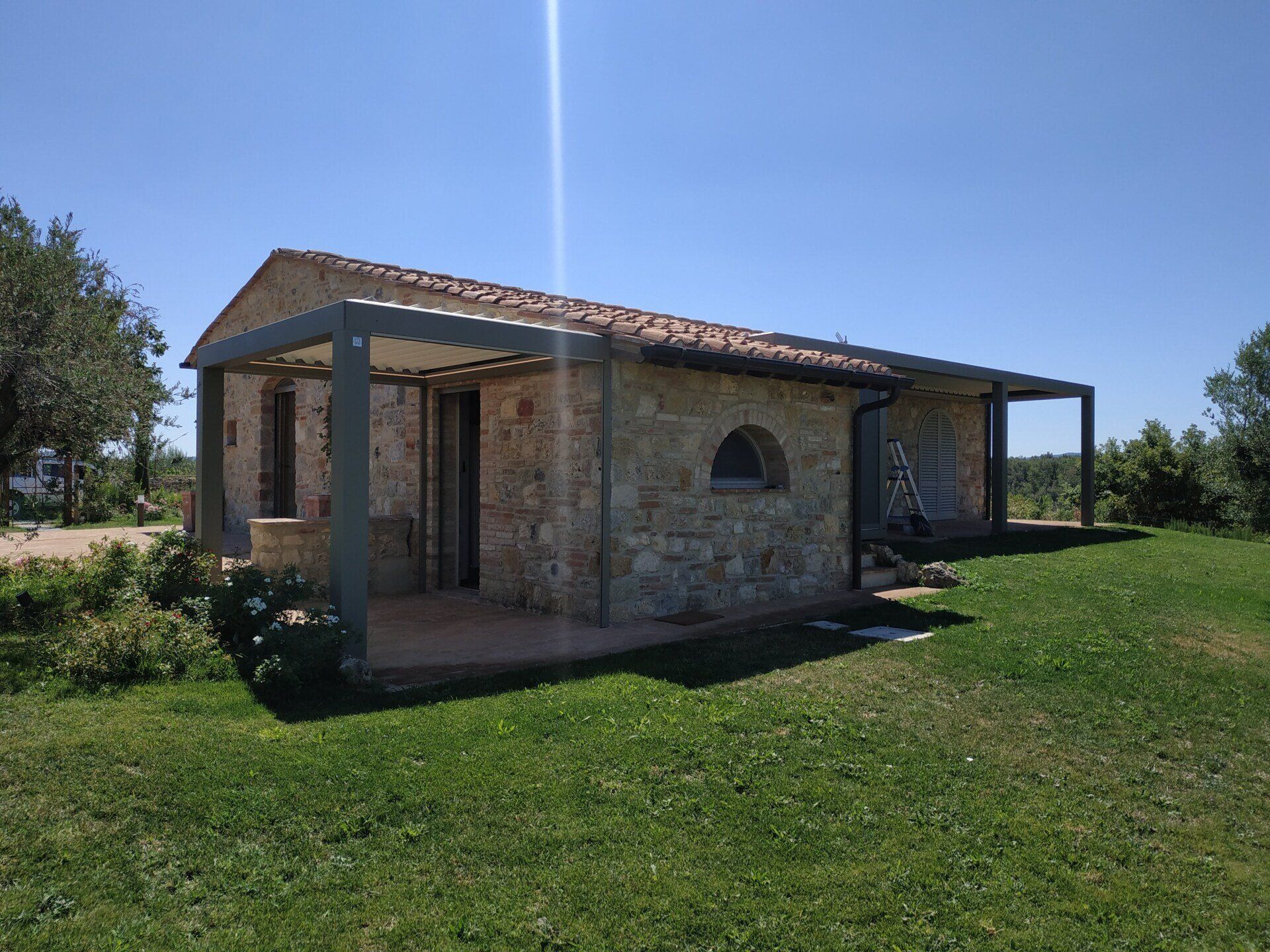Casa in pietra con tetto di tegole rosse e portico, situata in un soleggiato cortile erboso sotto un cielo azzurro e limpido.
