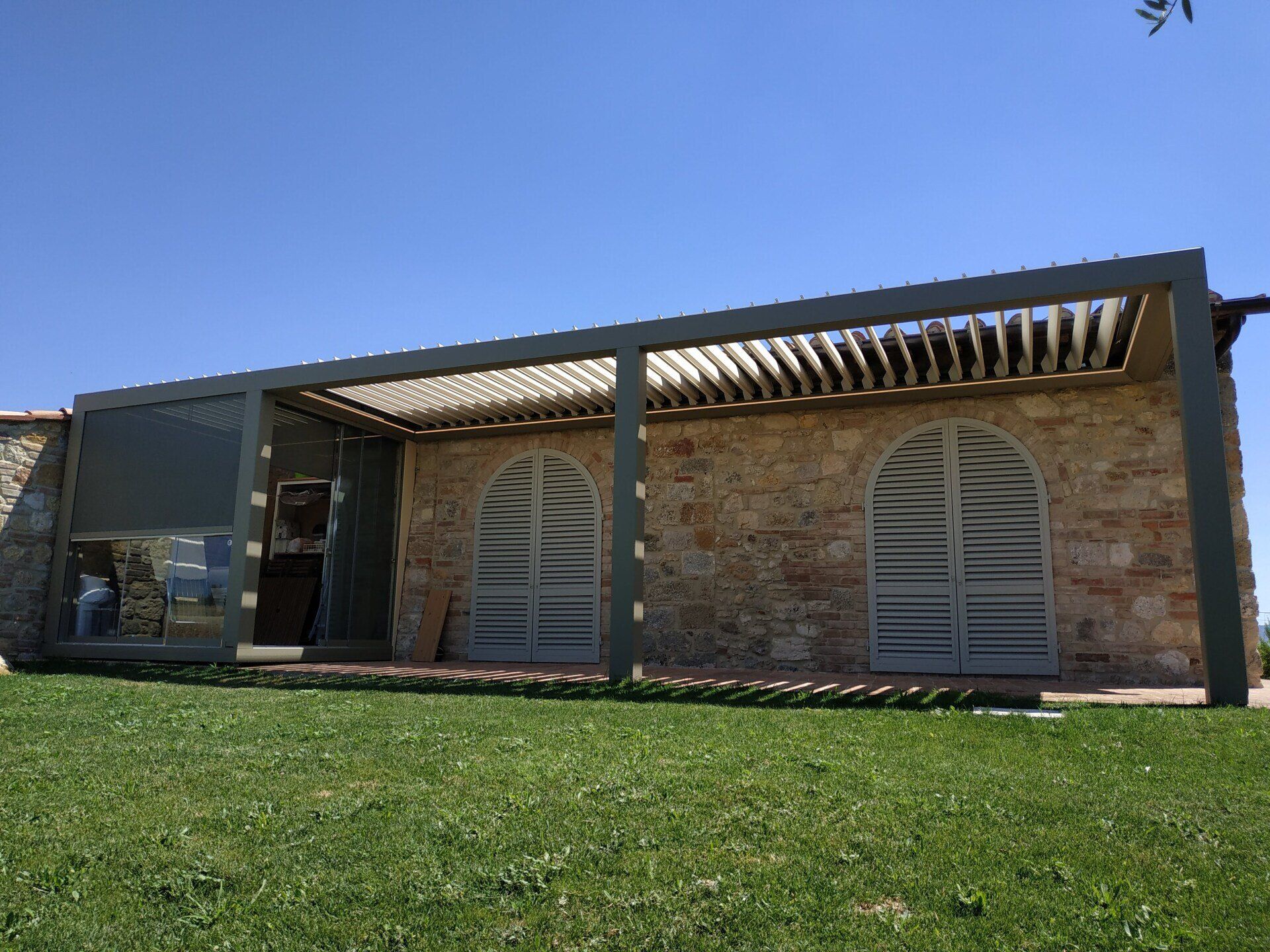 Casa in pietra con porte ad arco e pergolato, vista attraverso un prato verde sotto un cielo azzurro limpido.